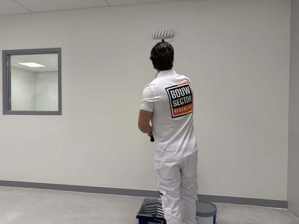 Schilder van Bouwsector Nederland rolt een witte wand, met verfbak en emmer op de vloer