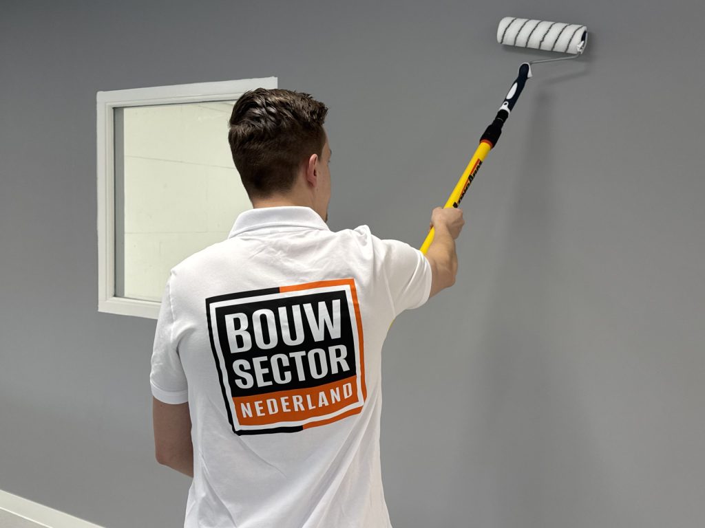 Schilder verft een grijze wand naast een wit gelakt raamkozijn
