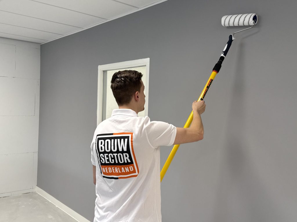 Vakman met verlengsteel saust een grijze wand in een lege ruimte met systeemplafond