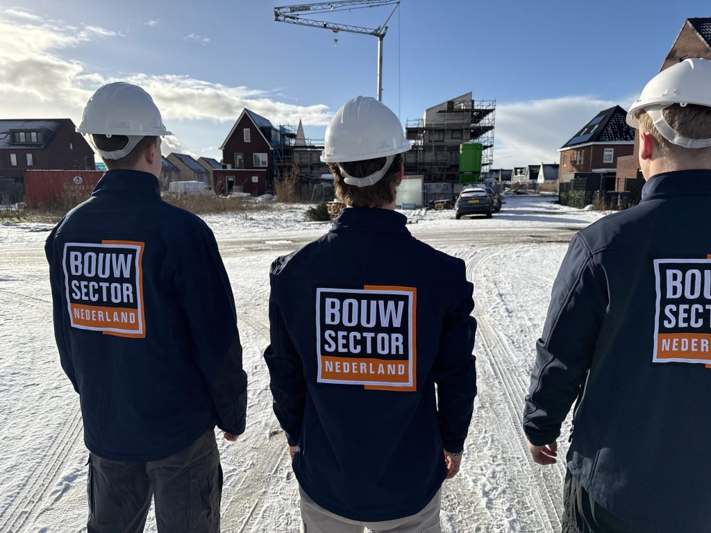 Drie vakmensen van Bouwsector Nederland in donkere werkjas en witte helm kijken naar huizen in aanbouw